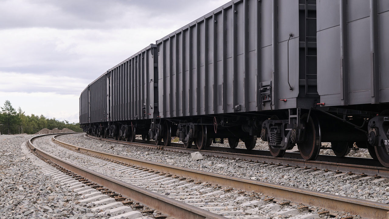 A row of black freight train cars on curved tracks surrounded by gravel and trees under a cloudy sky