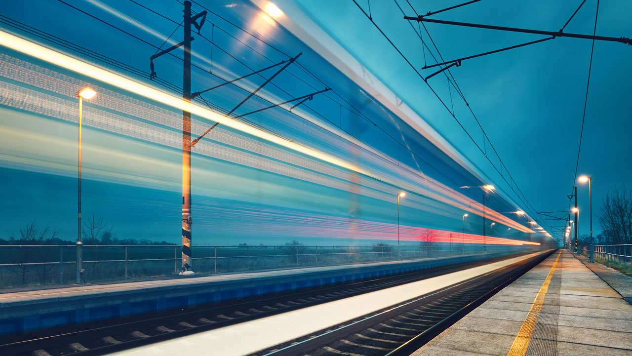 A blurred image of a train moving past a station, with lights and poles visible on a cloudy evening
